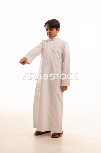 Gestures indicating this place (here), gestures to determine a specific location, portrait of a Saudi Arabian Gulf boy wearing traditional thobe and pointing with his index finger at something with an angry expression, full-length body image, white background