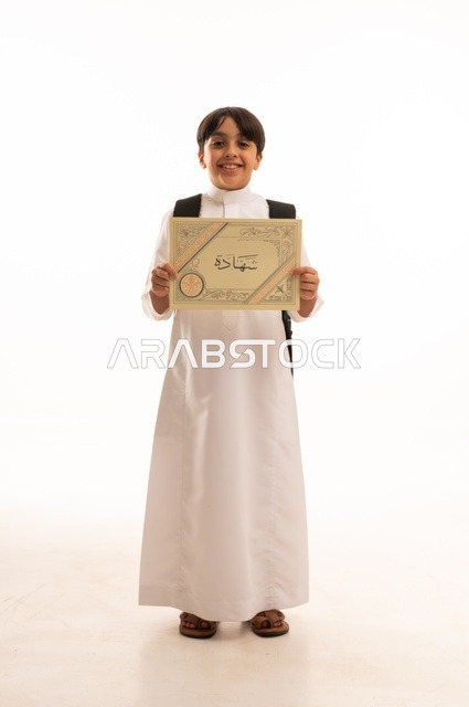 بالنجاح وانتهاء العام الدراسي، مفهوم التفوق والتخرج من المدرسة, النظر الى الكاميرا بايماءات الفخر والسعاده, التعليم الأكاديمي في السعوديه, بورتريه  لطالب عربي سعودي خليجي مبتسم يرتدي الثوب التقليدي يمسك بيده الشهادة، الجسم بالطول الكامل، خلفية بيضاء