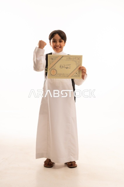 الاحتفال بالنجاح وانتهاء العام الدراسي، مفهوم التفوق والتخرج من المدرسة, النظر الى الكاميرا بايماءات الفخر والسعاده, التعليم الأكاديمي في السعوديه, بورتريه  لطالب عربي سعودي خليجي مبتسم يرتدي الثوب التقليدي يمسك بيده الشهادة، الجسم بالطول الكامل، خلفية بيضاء