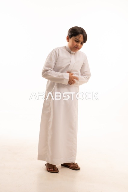 الإحساس بالمغص و التعب والإرهاق, طلب المساعده للذهاب الى الطبيب، عسر الهضم وحرقة في المعدة، بورتريه لولد عربي خليجي سعودي يرتدي الثوب التقليدي يضع يديه على معدته بإيماءات الشعور بألم في البطن, صورة الجسم بالكامل، خلفية بيضاء