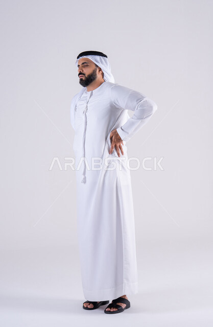 The need for treatment and a visit to the doctor, bone and joint pain, feeling tired and exhausted, a side portrait of an Arab Gulf Emirati man wearing a kandura and ghutra, placing his hands on his back with an expression of pain.