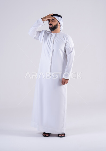 Feeling sick and having a headache, migraine pain, need for treatment and help, portrait of an Arab Gulf Emirati man wearing a kandura and ghutra, placing his hands on his head with gestures of annoyance and fatigue.