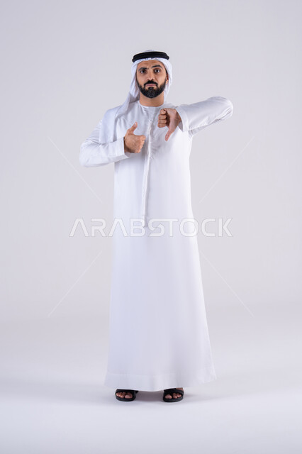 Confusion and hesitation about something. Portrait of an Arab Gulf Emirati man wearing a kandura and ghutra with hand gestures indicating disagreement and approval, dislike and admiration, rejection and acceptance, thumbs up and down.