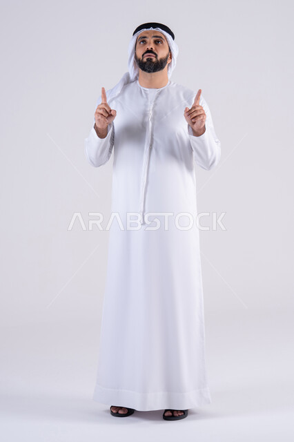 Expressions specifying this place here. Portrait of an Arab Gulf Emirati man wearing a kandura and ghutra, pointing upwards with his index finger at something, looking at the camera with gestures of joy and pleasure, specifying a specific location.