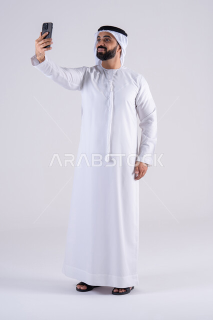 اجراء مكالمه فيديو ، استخدام التقنيات والتكنولوجيا الحديثة بالحياة اليومية ،الدردشة والتحدث مع الأصدقاء،تصفح مواقع وتطبيقات الانترنت، بورتريه لرجل عربي خليجي اماراتي يرتدي الكندوره والغتره، يمسك الجوال بيده