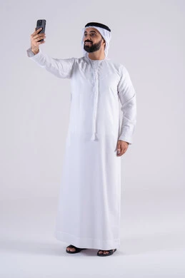 اجراء مكالمه فيديو ، استخدام التقنيات والتكنولوجيا الحديثة بالحياة اليومية ،الدردشة والتحدث مع الأصدقاء،تصفح مواقع وتطبيقات الانترنت، بورتريه لرجل عربي خليجي اماراتي يرتدي الكندوره والغتره، يمسك الجوال بيده