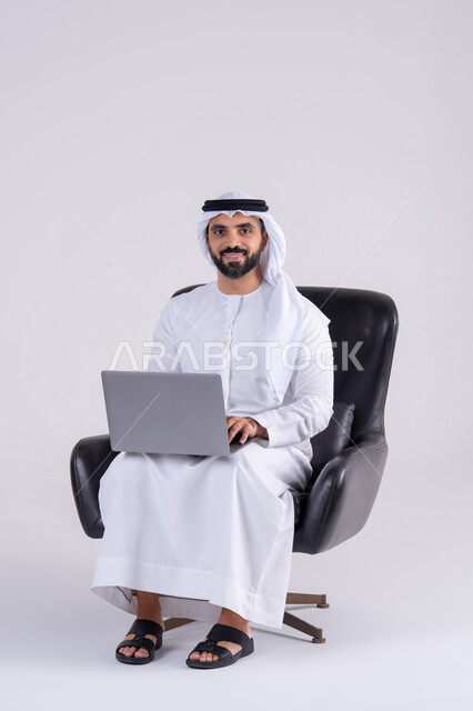 استخدم التقنيات الحديثة في الأعمال الإدارية ، انجاز مهام العمل عن بعد،اجراء مكالمات الفيديو، بورتريه لرجل عربي خليجي اماراتي يرتدي الكندوره والغتره، يجلس على كرسي ويعمل على جهاز الحاسب المحمول ، دمج التكنولوجيا المتطوره فى الحياه اليوميه