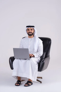 استخدم التقنيات الحديثة في الأعمال الإدارية ، انجاز مهام العمل عن بعد،اجراء مكالمات الفيديو، بورتريه لرجل عربي خليجي اماراتي يرتدي الكندوره والغتره، يجلس على كرسي ويعمل على جهاز الحاسب المحمول ، دمج التكنولوجيا المتطوره فى الحياه اليوميه