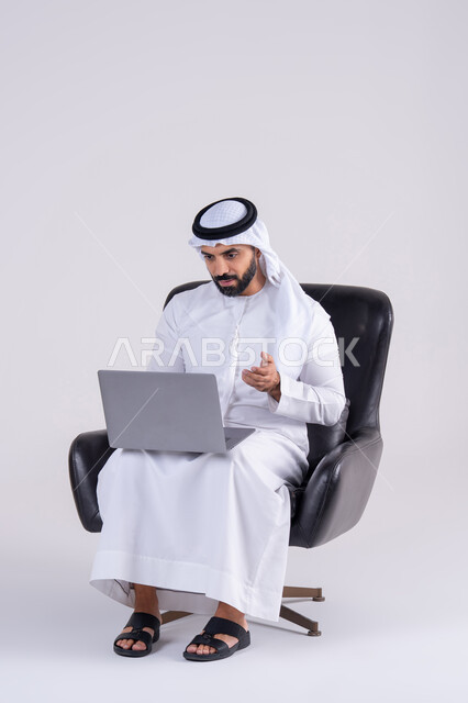 Completing work tasks remotely, making video calls, using modern technologies in administrative work, portrait of an Arab Gulf Emirati man wearing a kandora and ghutra, sitting on a chair and working on a laptop, integrating advanced technology into daily life.