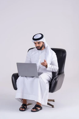 انجاز مهام العمل عن بعد،اجراء مكالمات الفيديو، استخدم التقنيات الحديثة في الأعمال الإدارية ، بورتريه لرجل عربي خليجي اماراتي يرتدي الكندوره والغتره، يجلس على كرسي ويعمل على جهاز الحاسب المحمول ، دمج التكنولوجيا المتطوره فى الحياه اليوميه