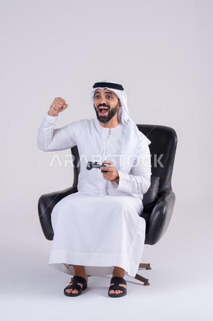 رفع قبضه اليد بايماءات الفوز والانتصار، مفهوم المتعة والتسلية، الاستمتاع بالتقنيات والتكنولوجيا الحديثة، قضاء أوقات ممتعة في اللعب بألعاب الفيديو،بورتريه لرجل عربي خليجي اماراتي يرتدي الكندوره والغتره يمسك بيده عصا التحكم