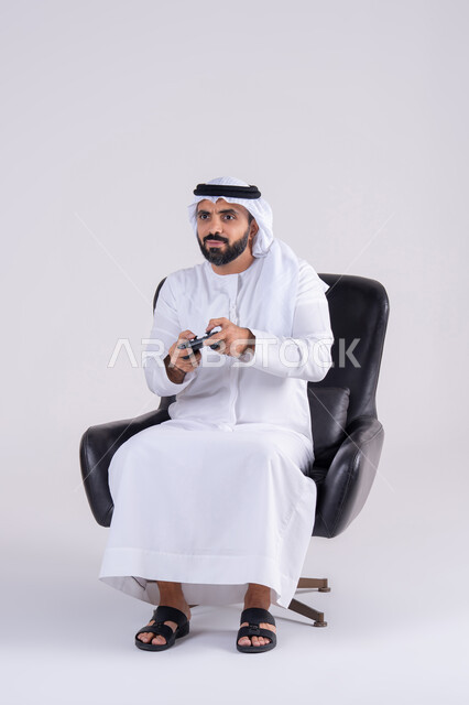 تعابير التركيز والاندماج ، رفع قبضه اليد بايماءات الفوز والانتصار، مفهوم المتعة والتسلية، قضاء أوقات ممتعة في اللعب بألعاب الفيديو،بورتريه لرجل عربي خليجي اماراتي يرتدي الكندوره والغتره يمسك بيده عصا التحكم