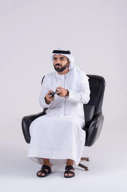 تعابير التركيز والاندماج ، رفع قبضه اليد بايماءات الفوز والانتصار، مفهوم المتعة والتسلية، قضاء أوقات ممتعة في اللعب بألعاب الفيديو،بورتريه لرجل عربي خليجي اماراتي يرتدي الكندوره والغتره يمسك بيده عصا التحكم
