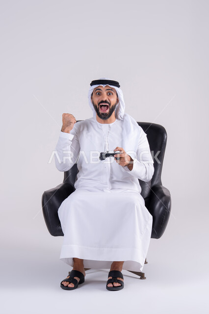 الاستمتاع بالتقنيات والتكنولوجيا الحديثة، رفع قبضه اليد بايماءات الفوز والانتصار، مفهوم المتعة والتسلية، قضاء أوقات ممتعة في اللعب بألعاب الفيديو،بورتريه لرجل عربي خليجي اماراتي يرتدي الكندوره والغتره يمسك بيده عصا التحكم