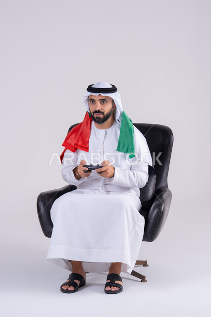 قضاء أوقات ممتعة في اللعب بألعاب الفيديو، مفهوم المتعة والتسلية، الاستمتاع بالتقنيات والتكنولوجيا الحديثة، رفع قبضة اليد بإيماءات الفوز والانتصار،بورتريه لرجل عربي خليجي اماراتي يرتدي الكندوره والغتره يمسك بيده عصا التحكم ويرتدي وشاح علم الامارات