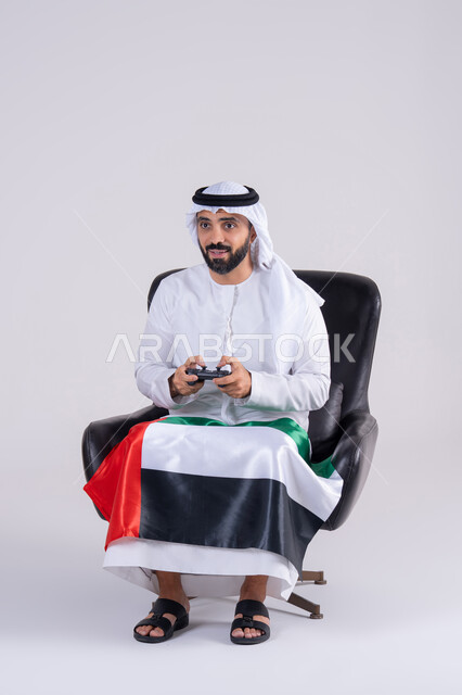 مفهوم المتعة والتسلية،قضاء أوقات ممتعة في اللعب بألعاب الفيديو،  الاستمتاع بالتقنيات والتكنولوجيا الحديثة، رفع قبضة اليد بإيماءات الفوز والانتصار،بورتريه لرجل عربي خليجي اماراتي يرتدي الكندوره والغتره يمسك بيده عصا التحكم ويرتدي وشاح علم الامارات