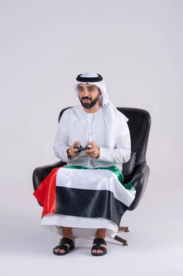The concept of fun and entertainment, having a good time playing video games, enjoying modern technology, raising a fist with a victory gesture, a portrait of an Arab Gulf Emirati man wearing a kandora and ghutra holding a joystick and wearing a UAE flag scarf.