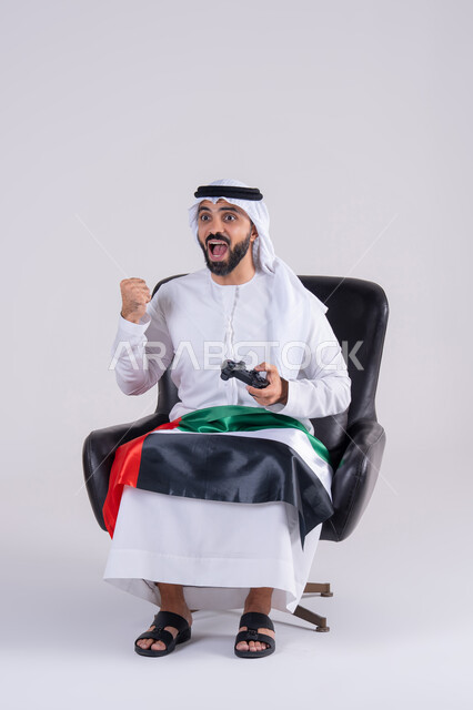 الاستمتاع بالتقنيات والتكنولوجيا الحديثة، مفهوم المتعة والتسلية،قضاء أوقات ممتعة في اللعب بألعاب الفيديو، رفع قبضة اليد بإيماءات الفوز والانتصار،بورتريه لرجل عربي خليجي اماراتي يرتدي الكندوره والغتره يمسك بيده عصا التحكم ويرتدي وشاح علم الامارات