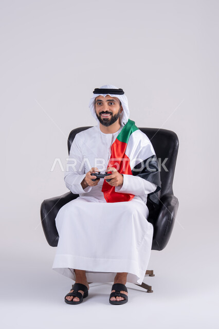 Having fun playing video games, raising a fist with victory gestures, enjoying modern technology, the concept of fun and entertainment, a portrait of an Arab Gulf Emirati man wearing a kandora and ghutra holding a joystick and wearing a UAE flag scarf.