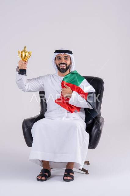 Following the match and supporting and cheering for the Arab national team. Portrait of an Arab Gulf Emirati man wearing a kandura and ghutra, holding a cup while watching an Arab League match at home. Facial gestures indicating victory. The concept of clubs and cheering.