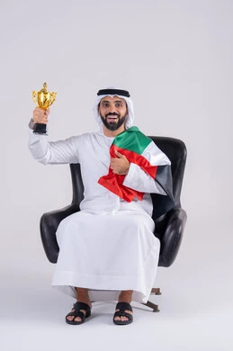 Following the match and supporting and cheering for the Arab national team. Portrait of an Arab Gulf Emirati man wearing a kandura and ghutra, holding a cup while watching an Arab League match at home. Facial gestures indicating victory. The concept of clubs and cheering.