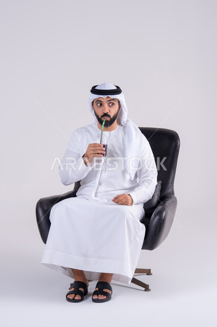 Gestures of wonder and amazement, having a good time at home, watching TV shows, home entertainment activities, drinking cold drinks, portrait of an Arab Gulf Emirati man wearing a kandura and ghutra, enjoying watching movies and TV series