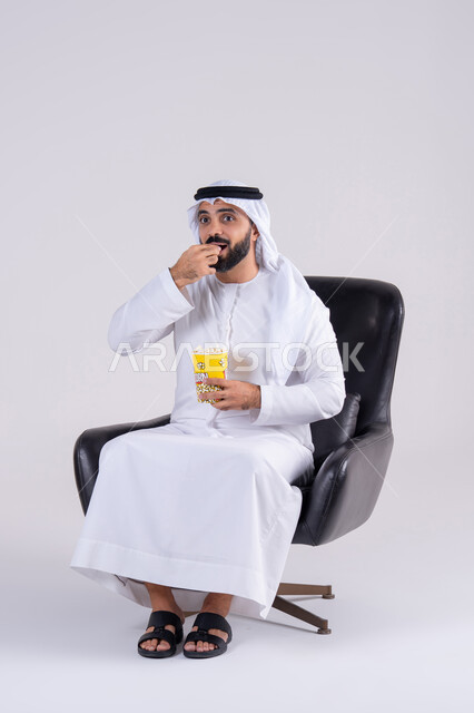 Having fun at home, enjoying popcorn, expressing surprise and amazement, and entertaining at home. A portrait of an Emirati Gulf Arab man wearing a kandura and ghutra, enjoying watching movies and TV series, and following TV shows.
