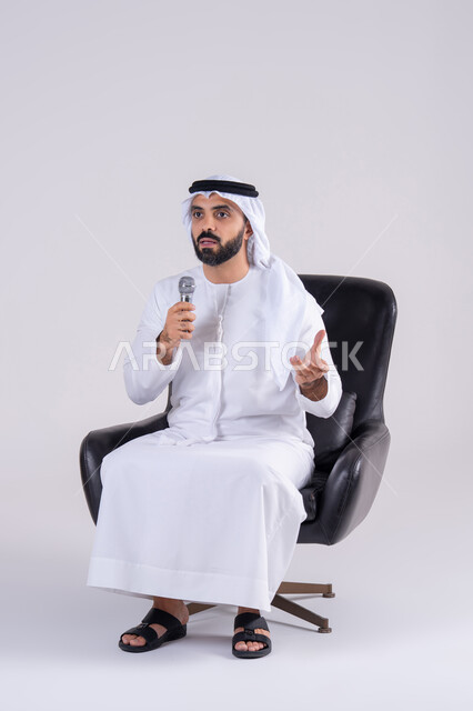 Recording a singing voice clip, presenting vocal performances, expressing immersion and interaction with music while singing, practicing singing talent, portrait of an Arab Gulf Emirati man wearing a kandura and ghutra, holding a microphone.