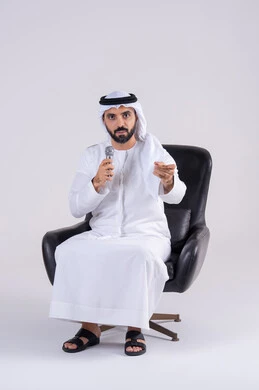 تسجيل مقطع صوتي غنائي، الاشاره باصبع السبابه ،ممارسة موهبة الغناء، بورتريه لرجل عربي خليجي اماراتي يرتدي الكندوره والغتره يمسك بيده المايكروفون ، تعابير الاندماج والتفاعل مع الموسيقي اثناء الغناء، تقديم العروض الصوتية