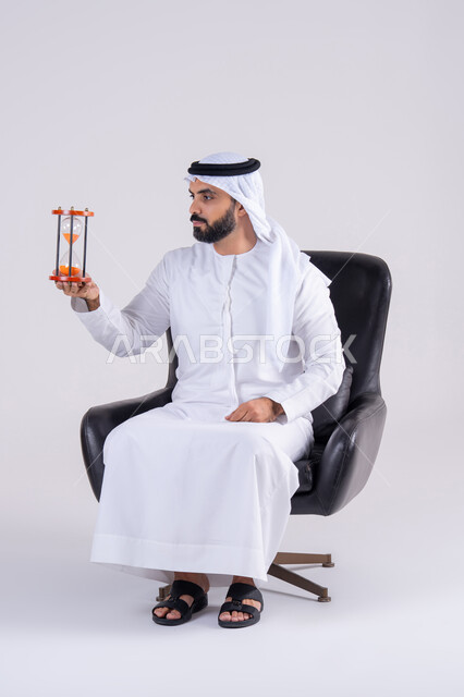 إدراك قيمة اللحظات الثمينة, مفهوم إدارة الأعمال وتنظيم الجدول الزمني, مراقبة توقيت الرمل, بورتريه لرجل عربي خليجي اماراتي يرتدي الكندوره والغتره، يمسك بيده الساعة الرملية الزجاجية (البنكام), النظر بإيماءات التركيز