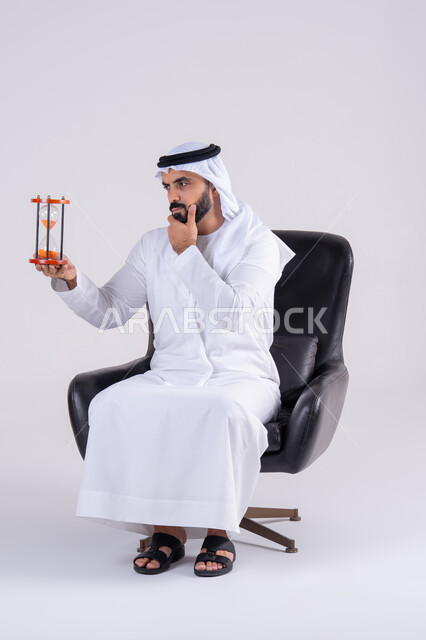 مفهوم إدارة الأعمال وتنظيم الجدول الزمني, مراقبة توقيت الرمل,إدراك قيمة اللحظات الثمينة,  بورتريه لرجل عربي خليجي اماراتي يرتدي الكندوره والغتره، يمسك بيده الساعة الرملية الزجاجية (البنكام), النظر بإيماءات التركيز