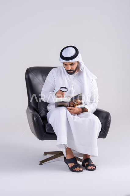 الفحص والاستكشاف والرؤية عن قرب، التدقيق باستخدام الأدوات البصرية،  مفهوم توضيح الصورة،بورتريه لرجل عربي خليجي اماراتي يرتدي الكندوره والغتره، يمسك بيده كتاب وينظر من خلال العدسة المكبرة بإيماءات التركيز