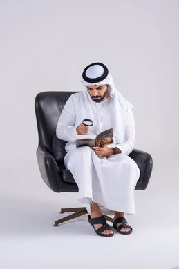 الفحص والاستكشاف والرؤية عن قرب، التدقيق باستخدام الأدوات البصرية،  مفهوم توضيح الصورة،بورتريه لرجل عربي خليجي اماراتي يرتدي الكندوره والغتره، يمسك بيده كتاب وينظر من خلال العدسة المكبرة بإيماءات التركيز