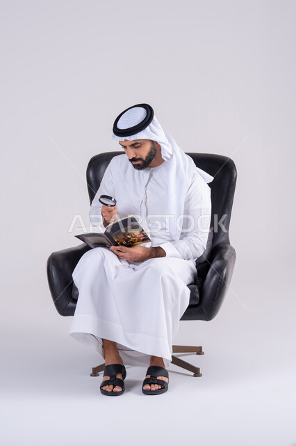 Scrutiny using optical instruments, examination, exploration, and close-up vision. The concept of image clarification. Portrait of an Arab Gulf Emirati man wearing a kandura and ghutra, holding a book and looking through a magnifying glass with focus gestures.
