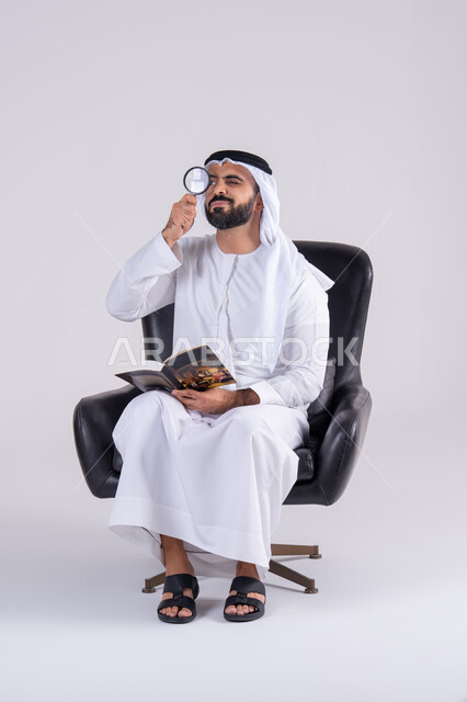 مفهوم توضيح الصورة، التدقيق باستخدام الأدوات البصرية، الفحص والاستكشاف والرؤية عن قرب، بورتريه لرجل عربي خليجي اماراتي يرتدي الكندوره والغتره، يمسك بيده كتاب وينظر من خلال العدسة المكبرة بإيماءات التركيز