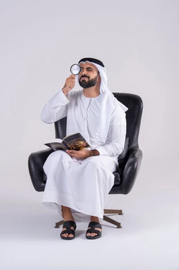 The concept of image clarification, scrutiny using optical tools, examination, exploration, and close-up. Portrait of an Arab Gulf Emirati man wearing a kandura and ghutra, holding a book and looking through a magnifying glass with focus gestures.