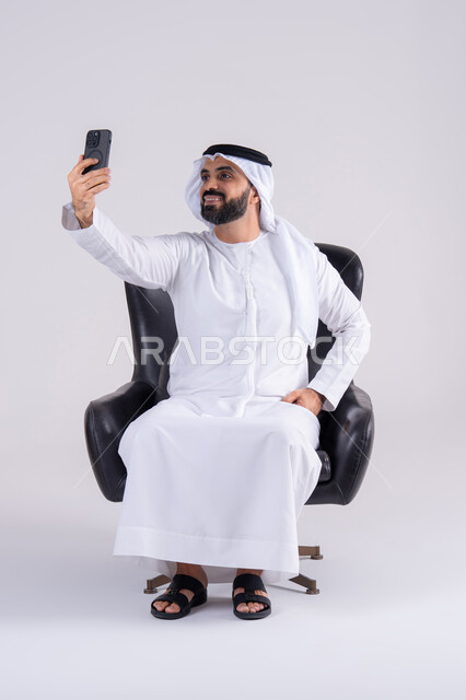 التقاط صور السيلفي ، عرض شاشه الهاتف المحمول ، الدردشة والتحدث مع الأصدقاء، بورتريه لرجل عربي خليجي اماراتي يرتدي الكندوره والغتره يمسك الجوال بيده، استخدام التقنيات والتكنولوجيا الحديثة بالحياة اليومية