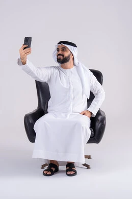 التقاط صور السيلفي ، عرض شاشه الهاتف المحمول ، الدردشة والتحدث مع الأصدقاء، بورتريه لرجل عربي خليجي اماراتي يرتدي الكندوره والغتره يمسك الجوال بيده، استخدام التقنيات والتكنولوجيا الحديثة بالحياة اليومية