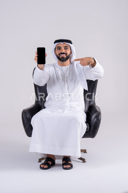 الاشاره الى شاشه الجوال ،التقاط صور السيلفي ،  الدردشة والتحدث مع الأصدقاء، بورتريه لرجل عربي خليجي اماراتي يرتدي الكندوره والغتره يمسك الهاتف المحمول بيده، استخدام التقنيات والتكنولوجيا الحديثة بالحياة اليومية