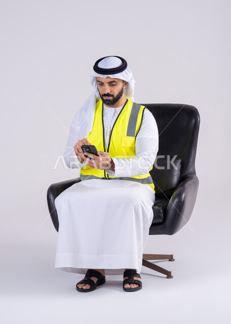 متابعه العمل والمهام،استخدام التقنيات الحديثة، الاشراف والرقابة على المشاريع الهندسية، بورتريه لرجل عربي خليجي اماراتي يرتدي الكندوره والغتره وستره الحمايه يجلس على كرسي ويحمل بيده الهاتف المحمول