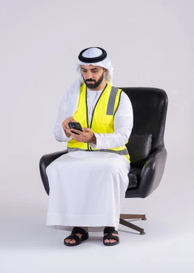 متابعه العمل والمهام،استخدام التقنيات الحديثة، الاشراف والرقابة على المشاريع الهندسية، بورتريه لرجل عربي خليجي اماراتي يرتدي الكندوره والغتره وستره الحمايه يجلس على كرسي ويحمل بيده الهاتف المحمول