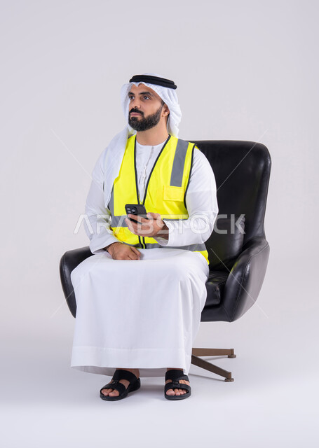 استخدام التقنيات الحديثة، متابعه العمل والمهام،الاشراف والرقابة على المشاريع الهندسية، بورتريه لرجل عربي خليجي اماراتي يرتدي الكندوره والغتره وستره الحمايه يجلس على كرسي ويحمل بيده الهاتف المحمول