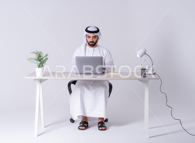 دمج التكنولوجيا المتطوره فى الحياه اليوميه، بورتريه لرجل عربي خليجي اماراتي يرتدي الكندوره والغتره يجلس على مكتب ويعمل على جهاز الحاسوب المحمول ،استخدم التقنيات الحديثة في الأعمال الإدارية ، التركيز والتفاني والعمل الجاد، التوظيف في المكاتب والشركات