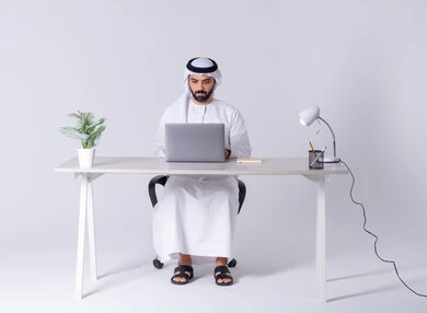 دمج التكنولوجيا المتطوره فى الحياه اليوميه، بورتريه لرجل عربي خليجي اماراتي يرتدي الكندوره والغتره يجلس على مكتب ويعمل على جهاز الحاسوب المحمول ،استخدم التقنيات الحديثة في الأعمال الإدارية ، التركيز والتفاني والعمل الجاد، التوظيف في المكاتب والشركات