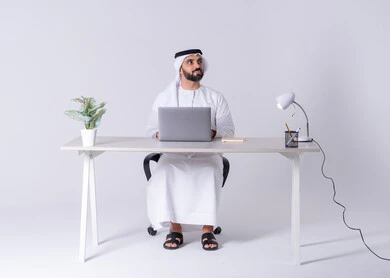 بورتريه لرجل عربي خليجي اماراتي يرتدي الكندوره والغتره يجلس على مكتب ويعمل على جهاز الحاسوب المحمول ،استخدم التقنيات الحديثة في الأعمال الإدارية ، التركيز والتفاني والعمل الجاد، التوظيف في المكاتب والشركات ،دمج التكنولوجيا المتطوره فى الحياه اليوميه