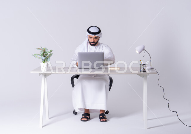 Use modern technologies in administrative work, focus, dedication and hard work, employment in offices and companies, portrait of an Emirati Gulf Arab man wearing a kandura and ghutra sitting at a desk and working on a laptop, integrating advanced technology into daily life