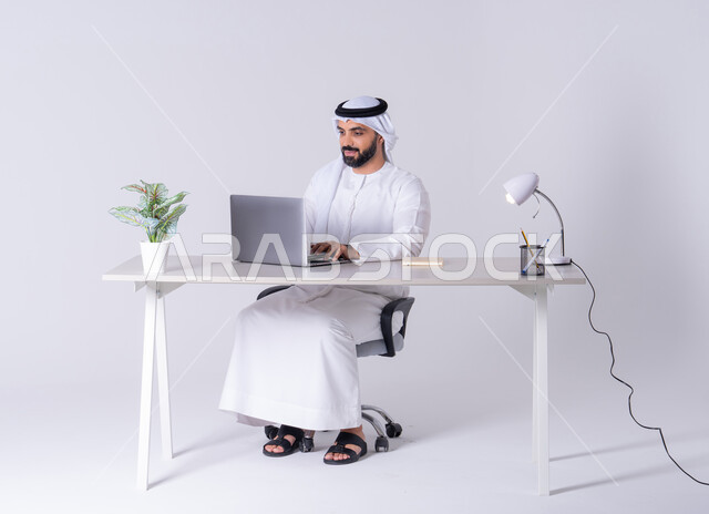 التركيز والتفاني والعمل الجاد، التوظيف في المكاتب والشركات ،استخدم التقنيات الحديثة في الأعمال الإدارية ، بورتريه لرجل عربي خليجي اماراتي يرتدي الكندوره والغتره يجلس على مكتب ويعمل على جهاز الحاسوب المحمول ،دمج التكنولوجيا المتطوره فى الحياه اليوميه
