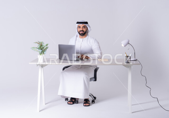 التوظيف في المكاتب والشركات ، التركيز والتفاني والعمل الجاد،استخدم التقنيات الحديثة في الأعمال الإدارية ، بورتريه لرجل عربي خليجي اماراتي يرتدي الكندوره والغتره يجلس على مكتب ويعمل على جهاز الحاسوب المحمول ،دمج التكنولوجيا المتطوره فى الحياه اليوميه
