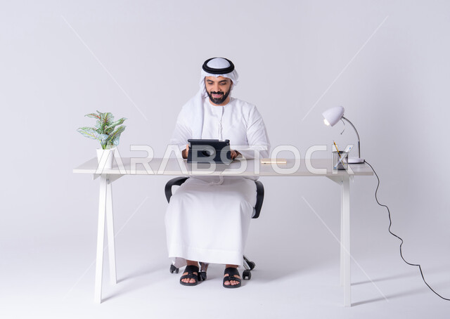 انجاز مهام العمل عن بعد ،اجراء مكالمه فيديو ،دمج التقنيات المتطوره فى الحياة اليومية، بورتريه لرجل عربي خليجي اماراتي يرتدي الكندوره والغتره يجلس عل مكتب ويمسك التابلت بيده