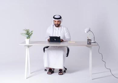 انجاز مهام العمل عن بعد ،اجراء مكالمه فيديو ،دمج التقنيات المتطوره فى الحياة اليومية، بورتريه لرجل عربي خليجي اماراتي يرتدي الكندوره والغتره يجلس عل مكتب ويمسك التابلت بيده